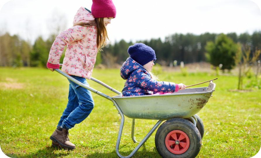 A girl pushing a wheelbarrow with a small child in it