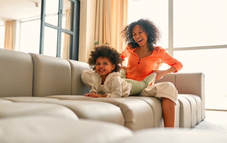 African American woman with her little daughter with curly fluffy hair sitting on the sofa and having fun in the living room at home.