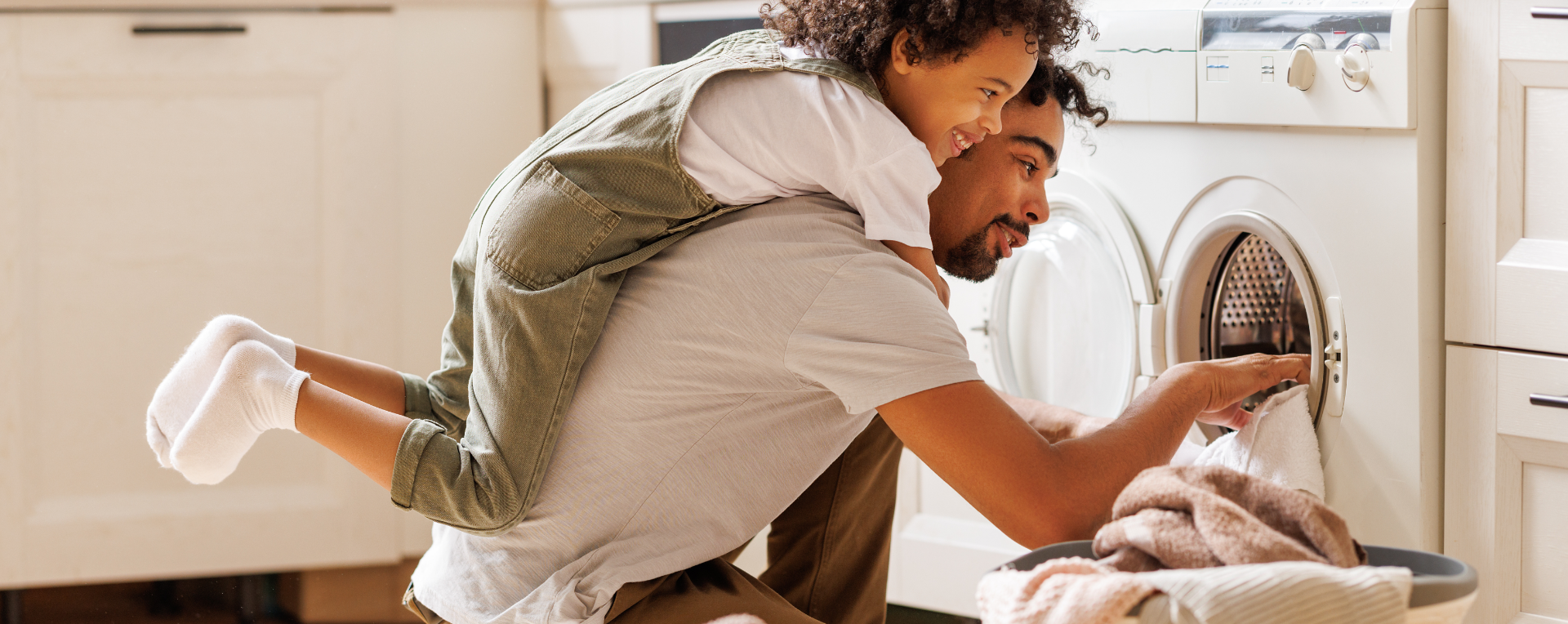 A man getting laundry out of a dryer with a young girl hanging on his back.