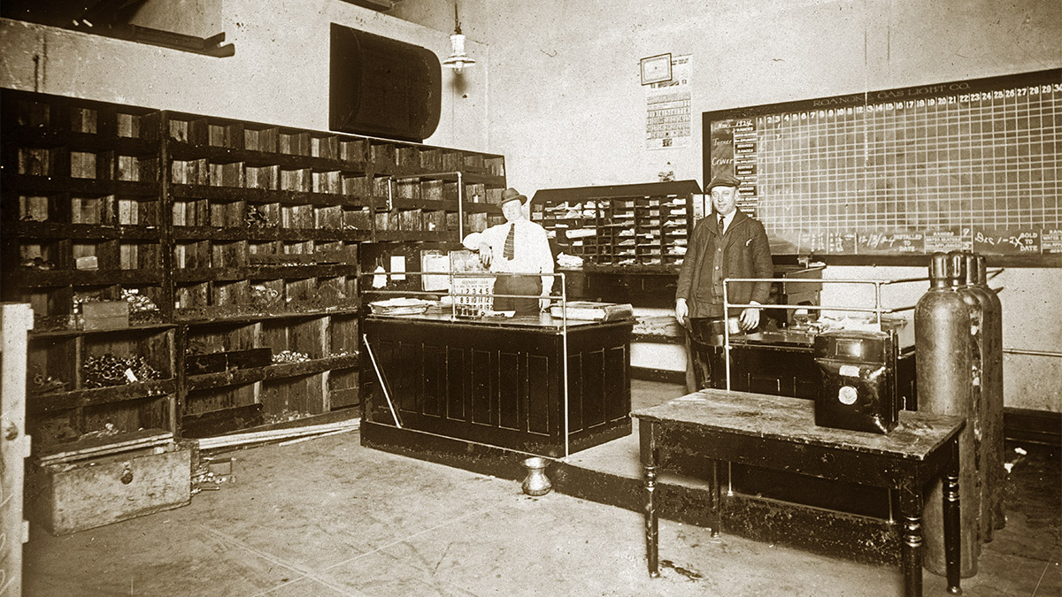 Two men standing at desks in an office with a wall lined with shelves