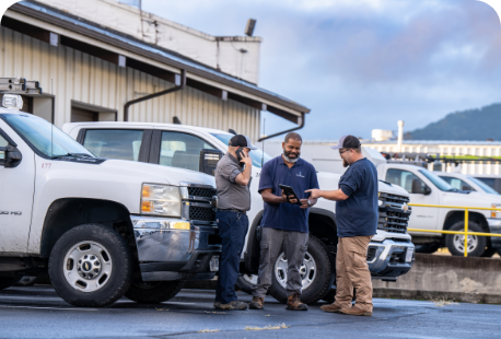 Three men standing in a parking loot in front of a work truck. One is on their phone while the other two are looking at a tablet.