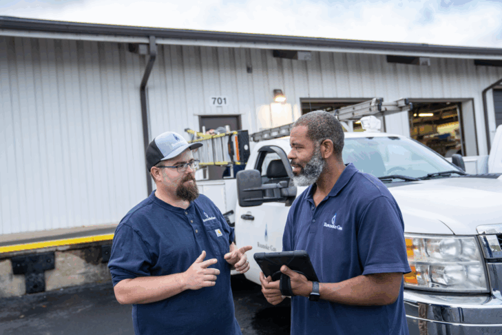 Two men standing in a parking lot talking with a work truck in the background behind them.