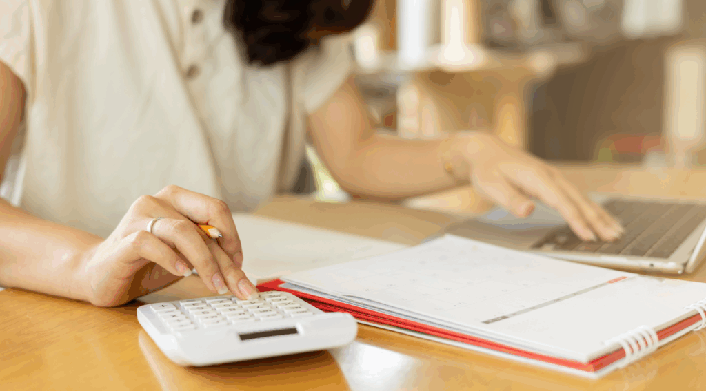 A person using a small calculator and laptop at a desk.