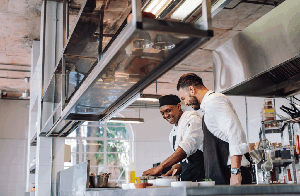 Two workers in a kitchen wearing aprons and preparing food.