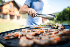 A grill covered with food being cooked. A person stands beside the grill holding a set of tongs.