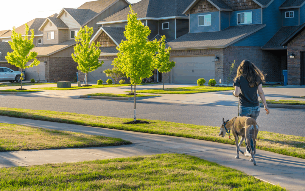 A woman walking a dog on a leash down a sidewalk in a residential area.
