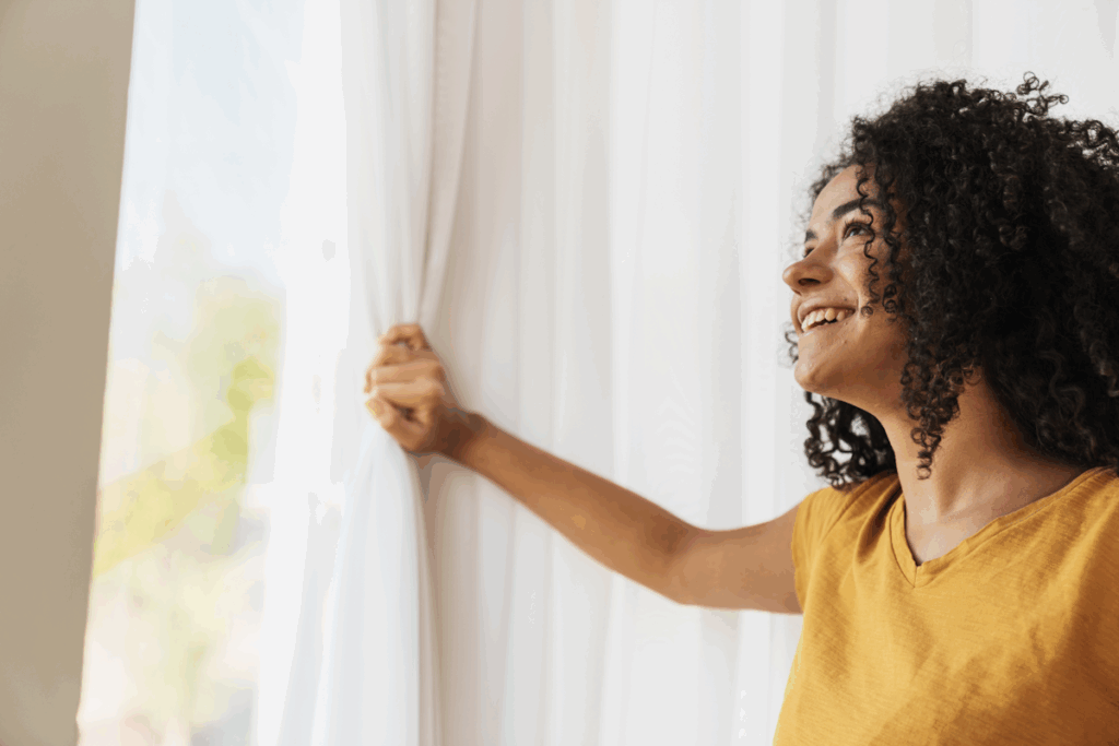 A woman pulling back a curtain and looking out on a sunny day.