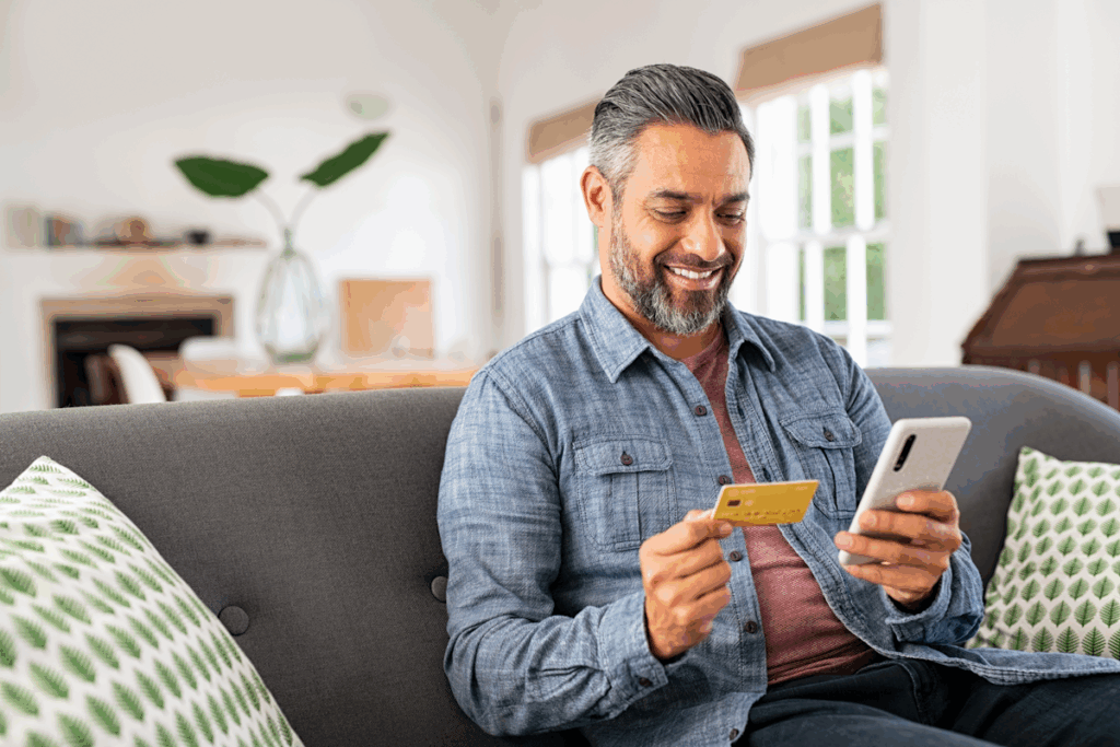 A man seated on a couch with a credit card in one hand and his smartphone in the other.