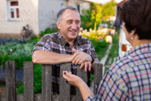 A man standing behind a picket fence with his are resting on it talking to a woman on the other side of the fence.