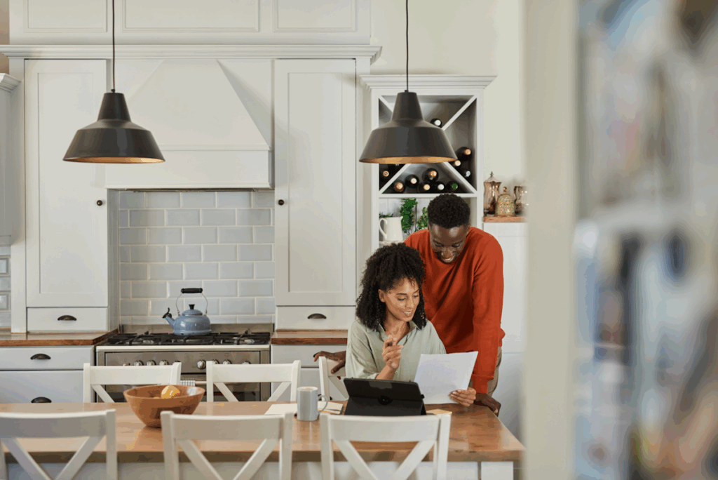 A woman seated at a table in a kitchen with a man standing behind her. They are both looking at a piece of paper she is holding.