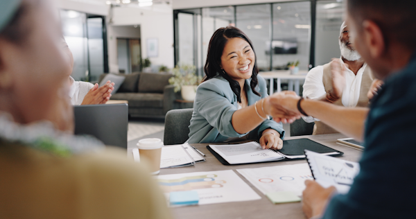 A group of people seated around a table. Two of them are reaching across the table and shaking hands.