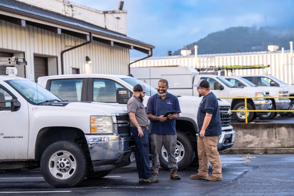 Three men standing in a parking lot in front of some work trucks. One man is holding a tablet.