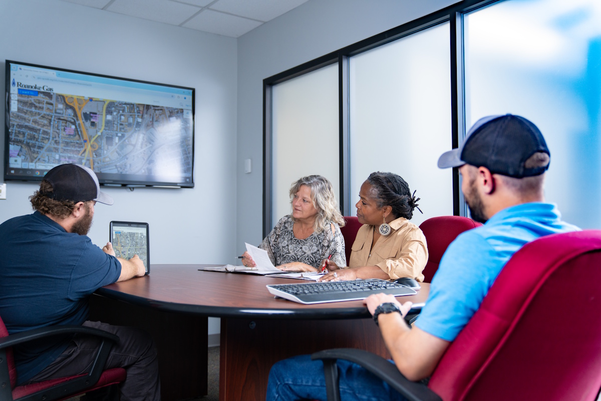 Two women and two men seated at a conference table. One of the men is showing an image on a tablet to the others.